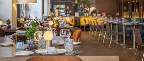 A restaurant with tables set for dining, featuring a modern interior with a bar area in the background.