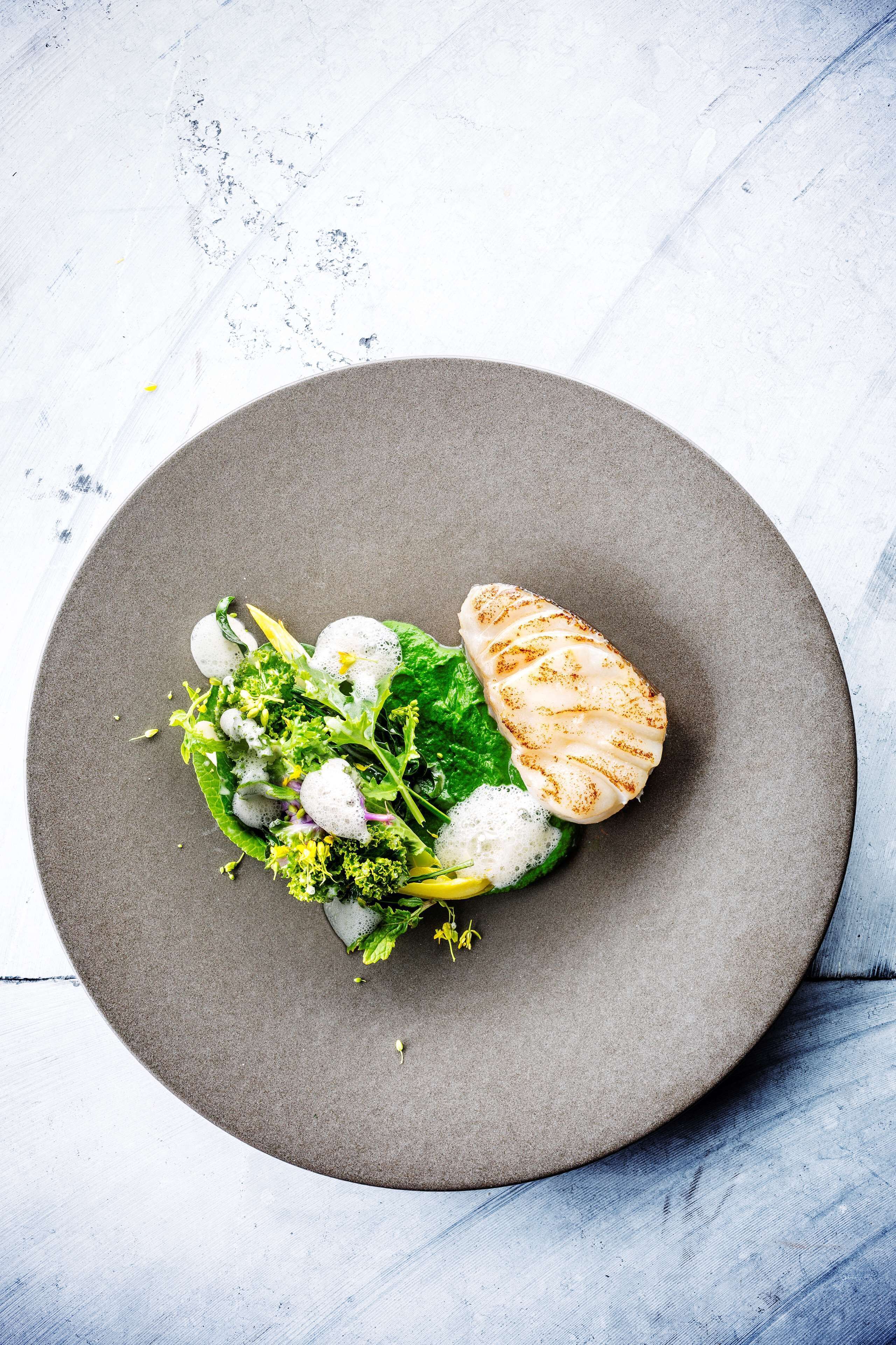 A plate with a grilled fish, greens, and a small salad on a round gray plate.