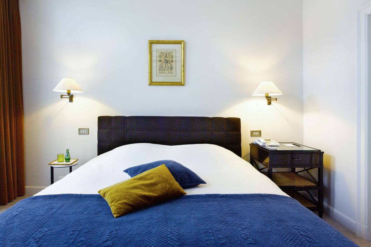 Een net opgemaakt bed met een blauwe quilt en een mosterdkleurig kussen, een nachtkastje met een lamp, en een lijst met een foto aan de muur.