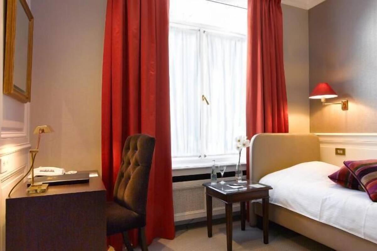 Hotelkamer met een bed, bureau, stoel, raam met gordijnen, een wandlamp en een lijst met een afbeelding.