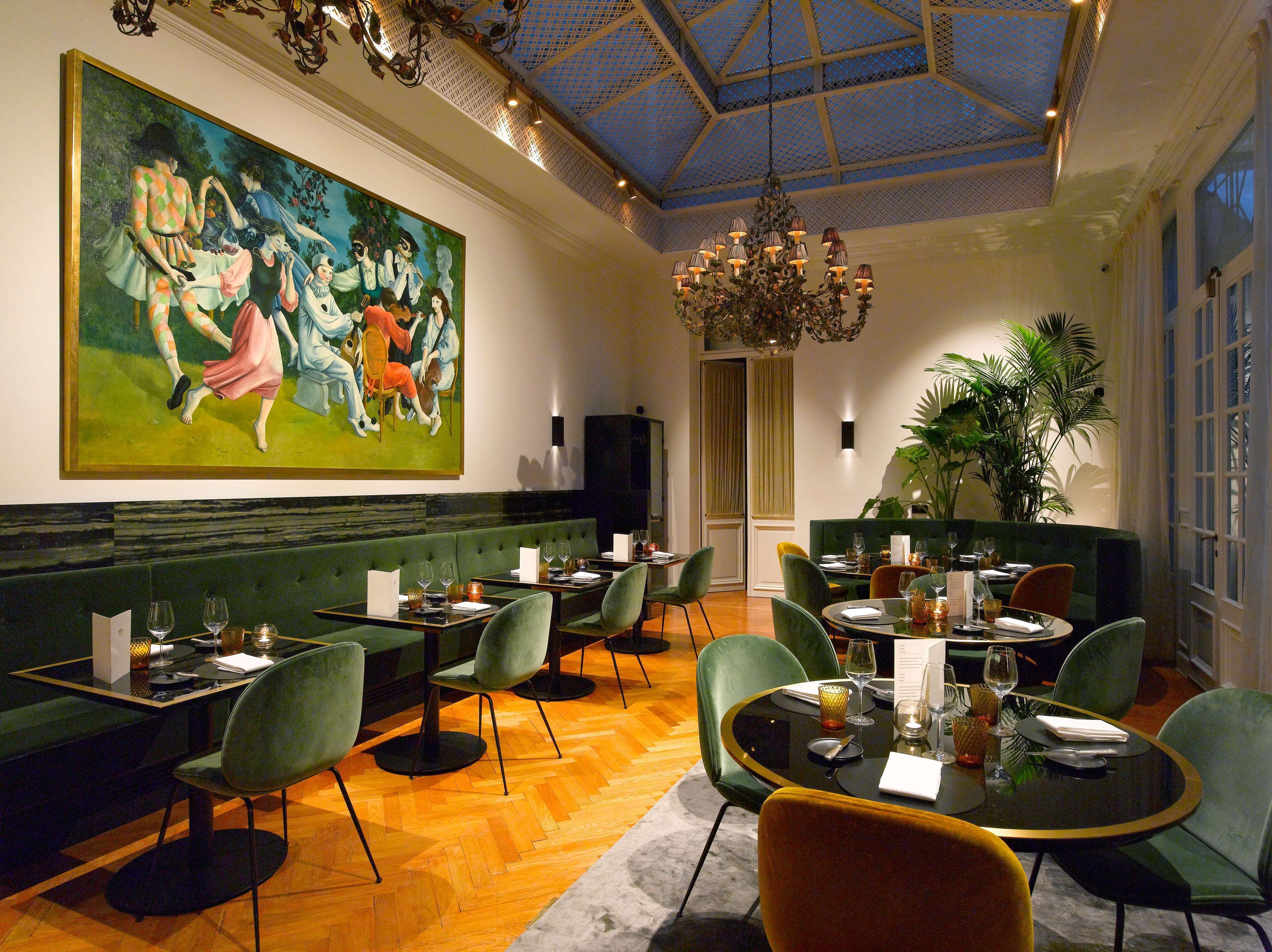 A dining area with green upholstered booths, round tables, and a large painting on the wall.