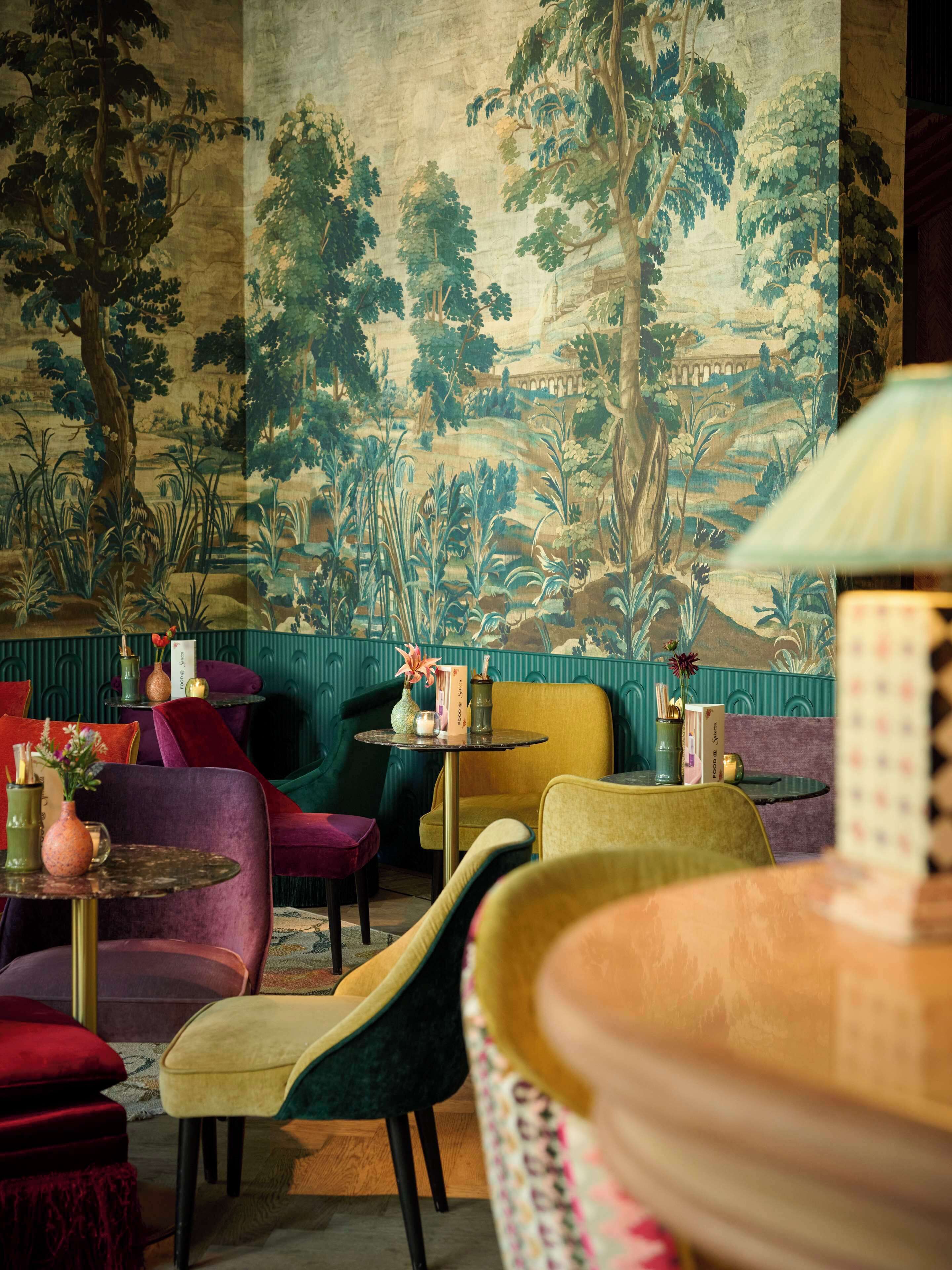 A room with a mural of a forest scene, plush seating, and a small table with a vase and candle.