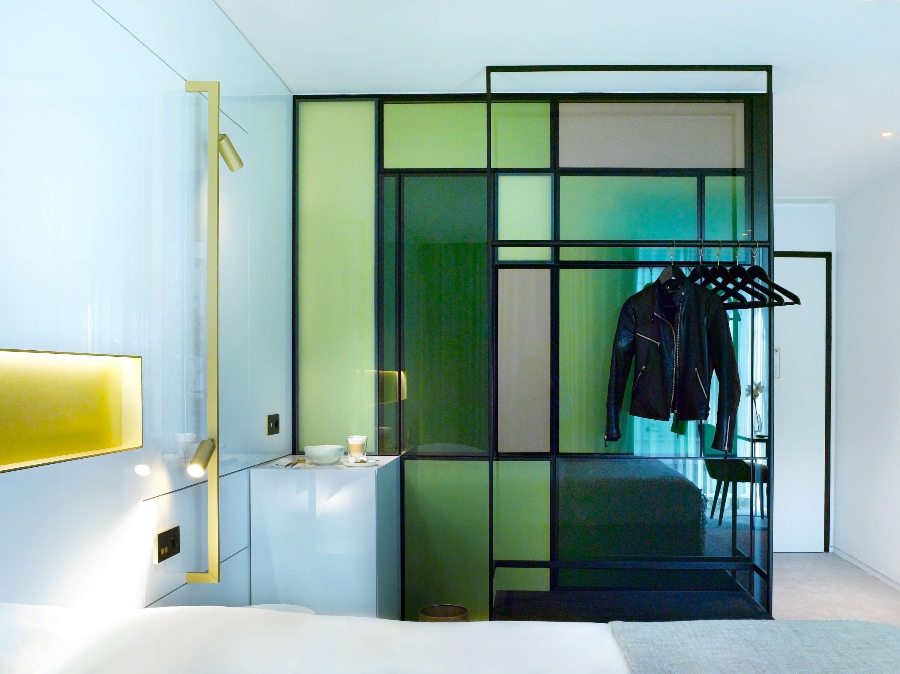 A modern hotel room with a bed, a bedside table with a lamp, a mirror, and a clothing rack with jackets.