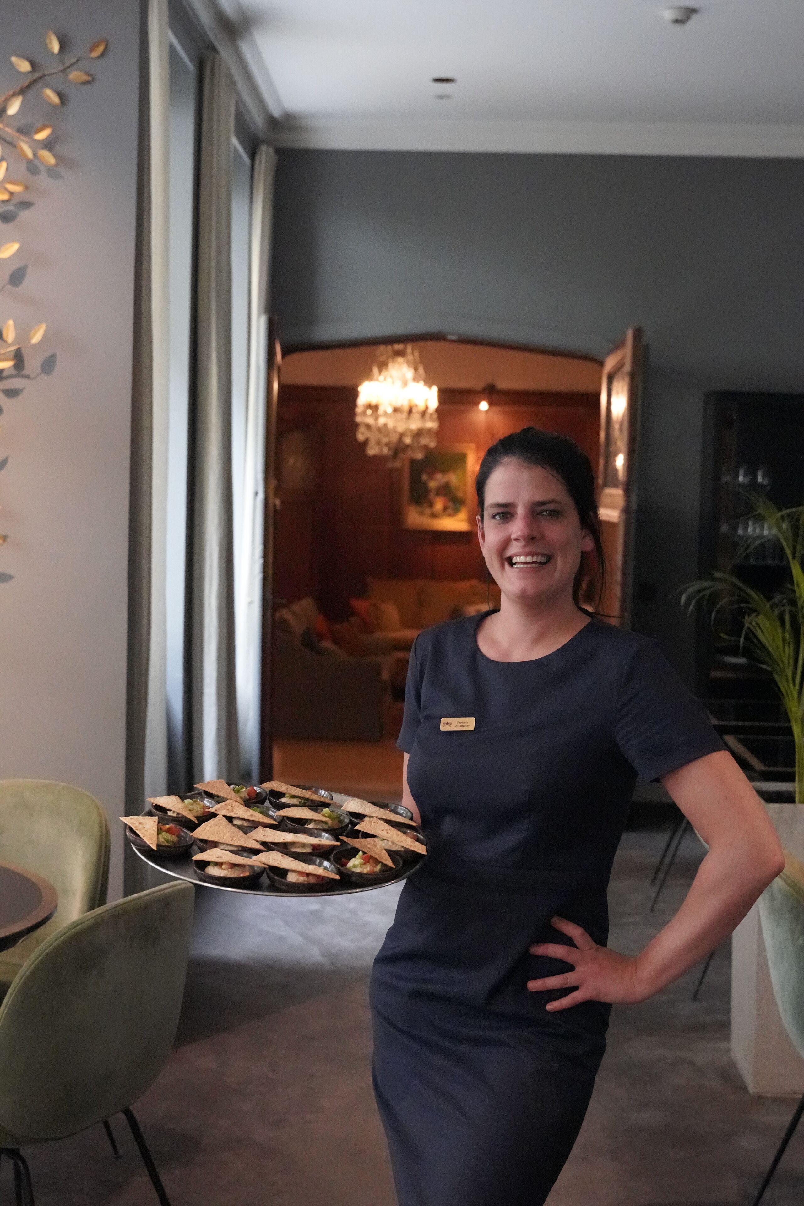 Mulher de vestido escuro carregando uma bandeja com comida em um hall moderno de hotel, com um candelabro e área de estar ao fundo.