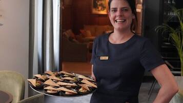 Mulher de vestido escuro carregando uma bandeja com comida em um hall moderno de hotel, com um candelabro e área de estar ao fundo.