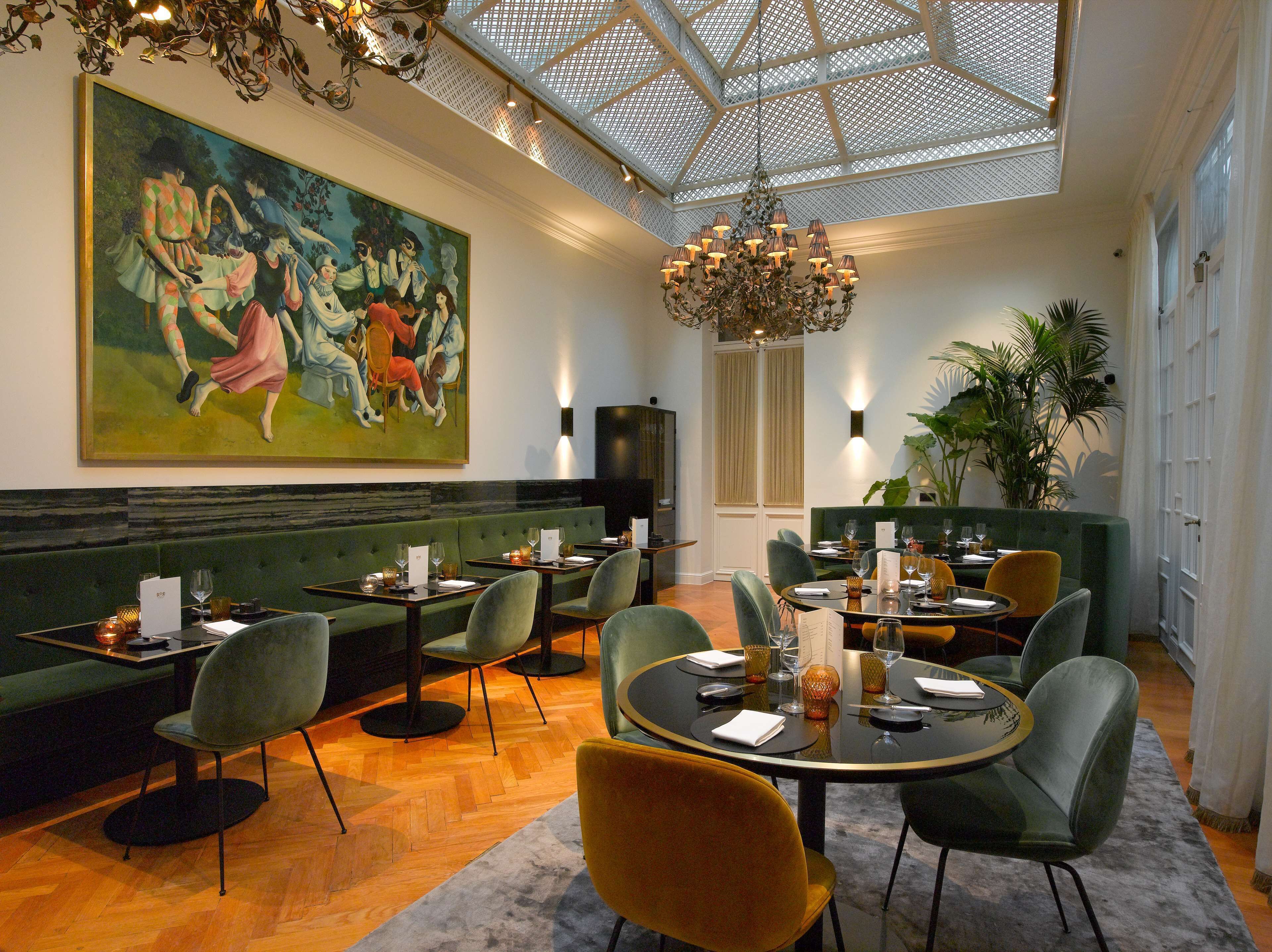 A dining area with green upholstered booths, round tables, and modern chairs. A large painting adorns the wall, and a chandelier hangs from the ceiling.