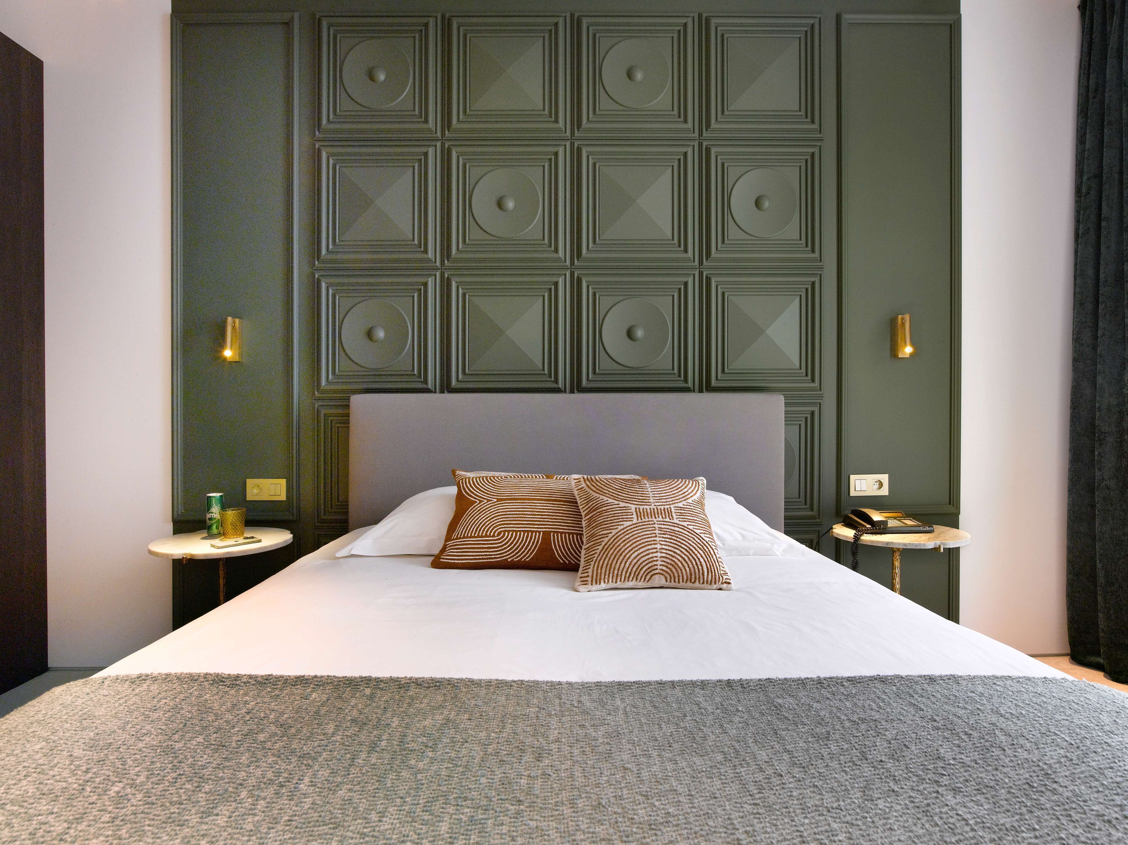 Una cama bien hecha con dos almohadones estampados, una mesita de noche con lámpara y una pared con diseño geométrico.