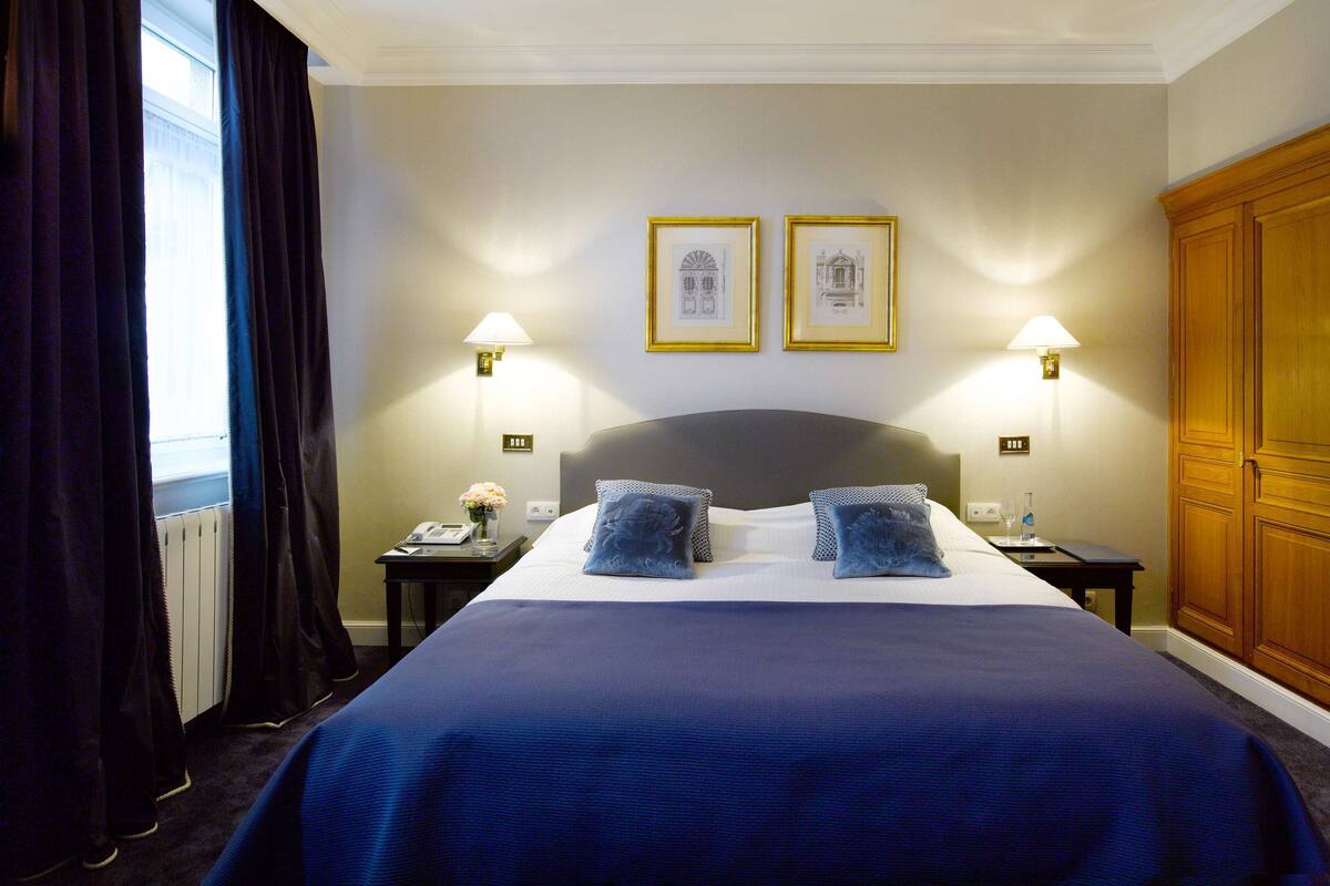 Een hotelkamer met een groot bed, twee bedlampjes, een nachtkastje met een telefoon en een bloevas, en een houten garderobe.
