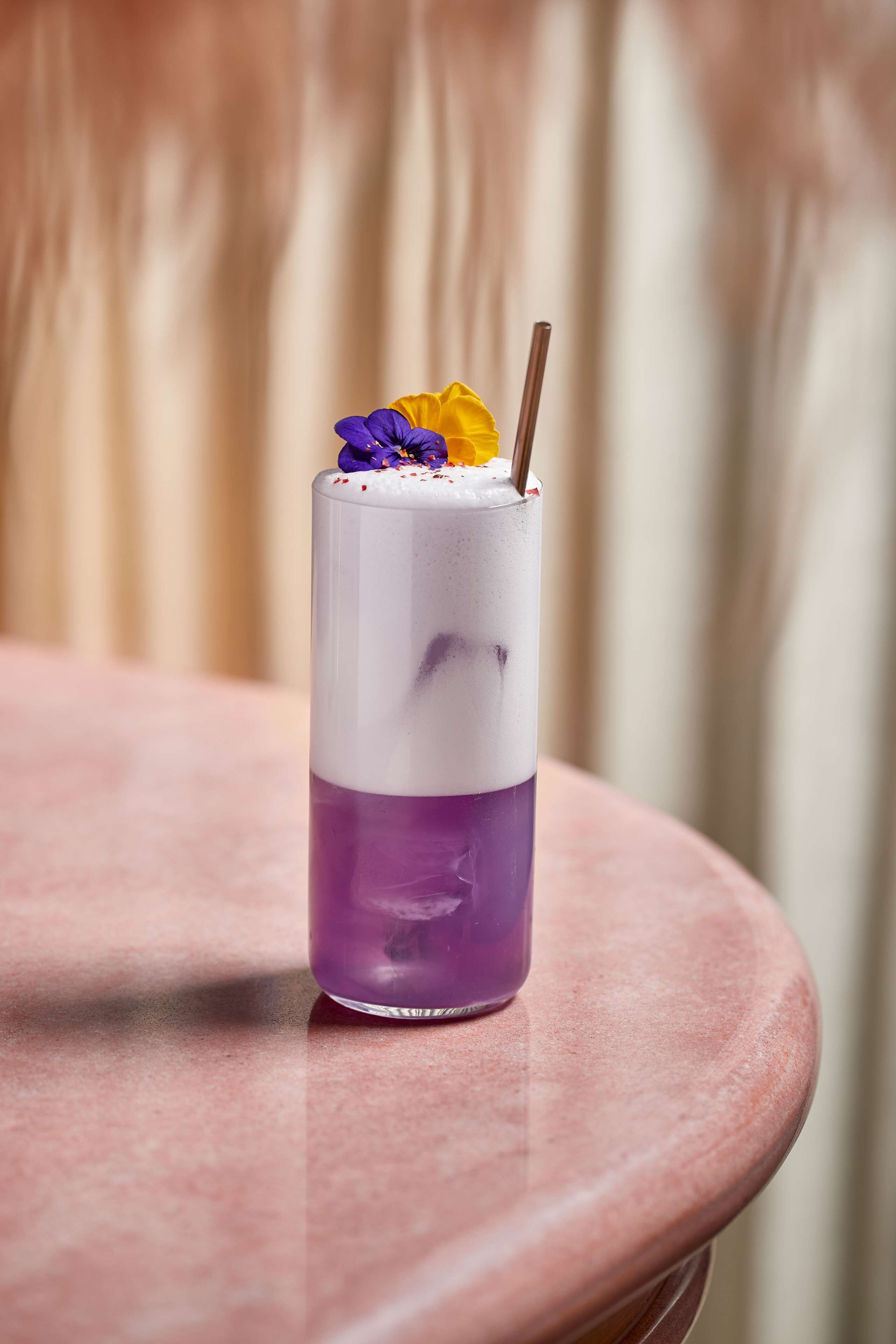 Un grand verre aux couleurs superposées, agrémenté d’une paille, posé sur une table ronde devant un rideau en arrière-plan.