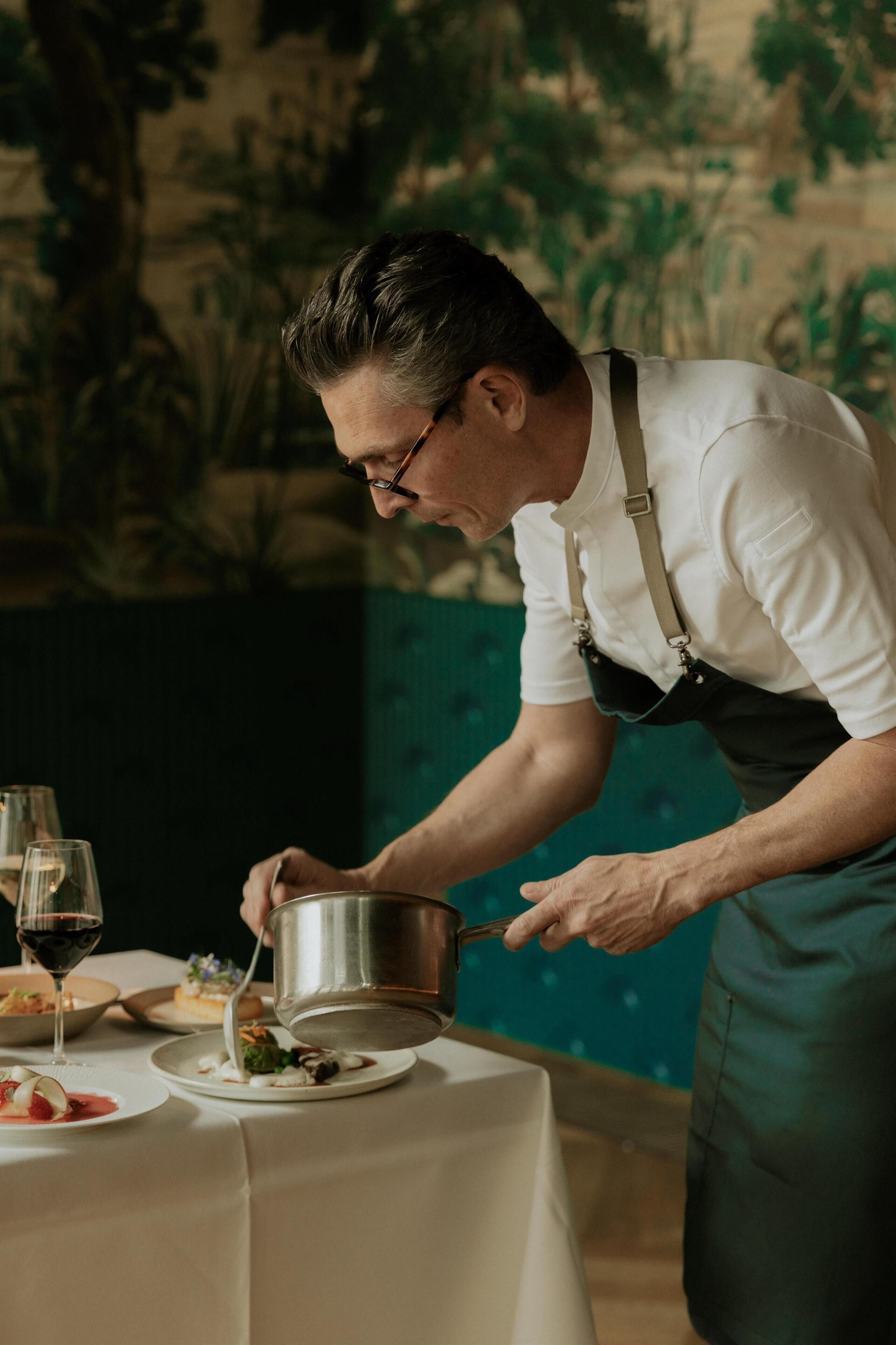 A person serving food at a table with a white tablecloth, wine glasses, and plates of food.