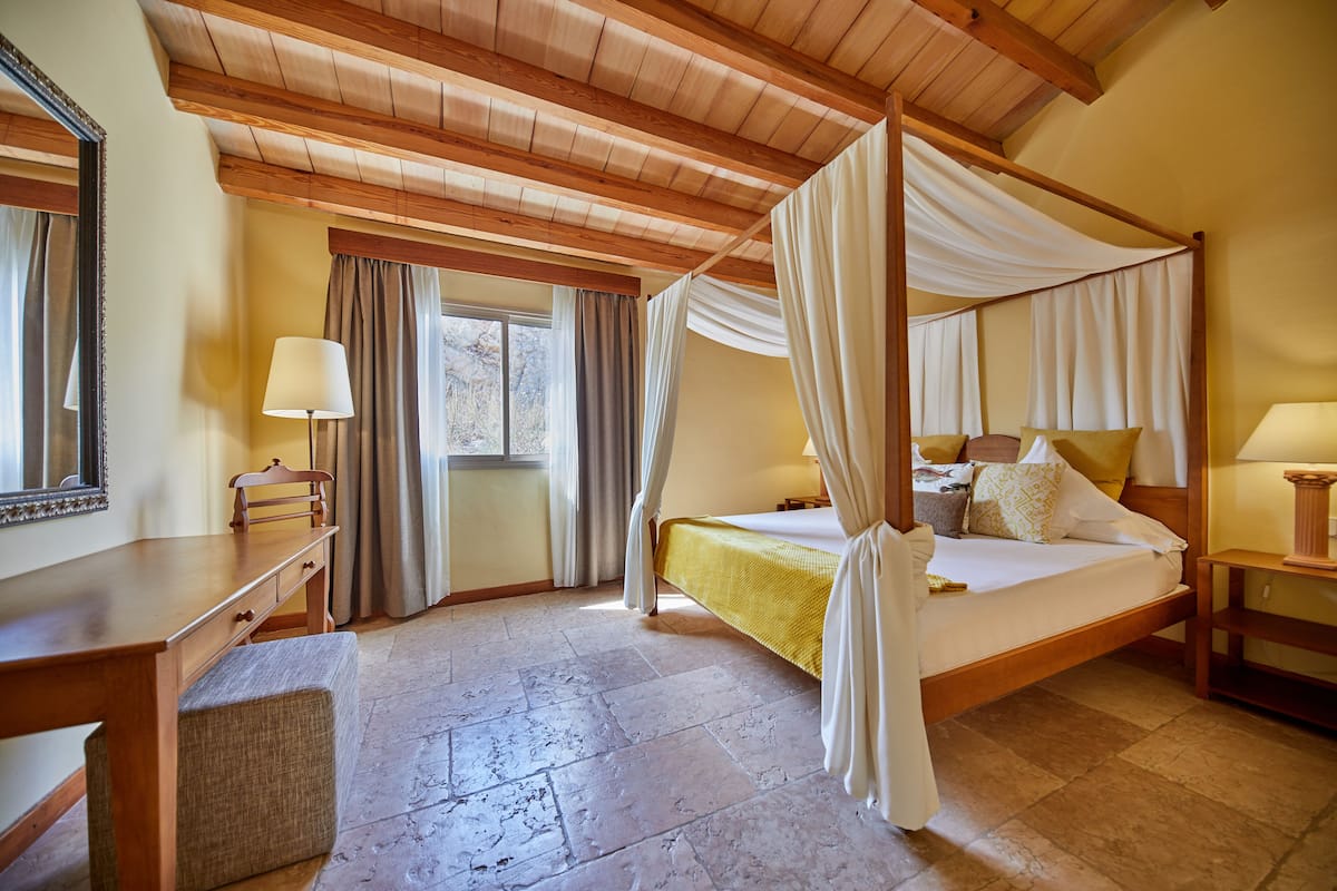 A four-poster bed with a canopy, a desk with a chair, a bedside table with a lamp, and a large mirror on the wall.
