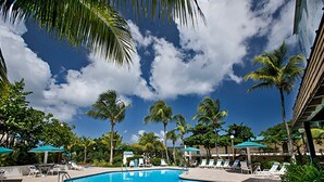 Una piscina con sillas de descanso y sombrillas, rodeada de palmeras y un cielo despejado.