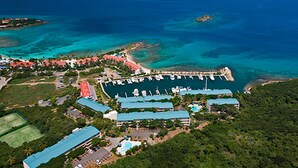 Vista aérea de un complejo turístico costero con varias edificaciones, un puerto deportivo y aguas azules cristalinas.