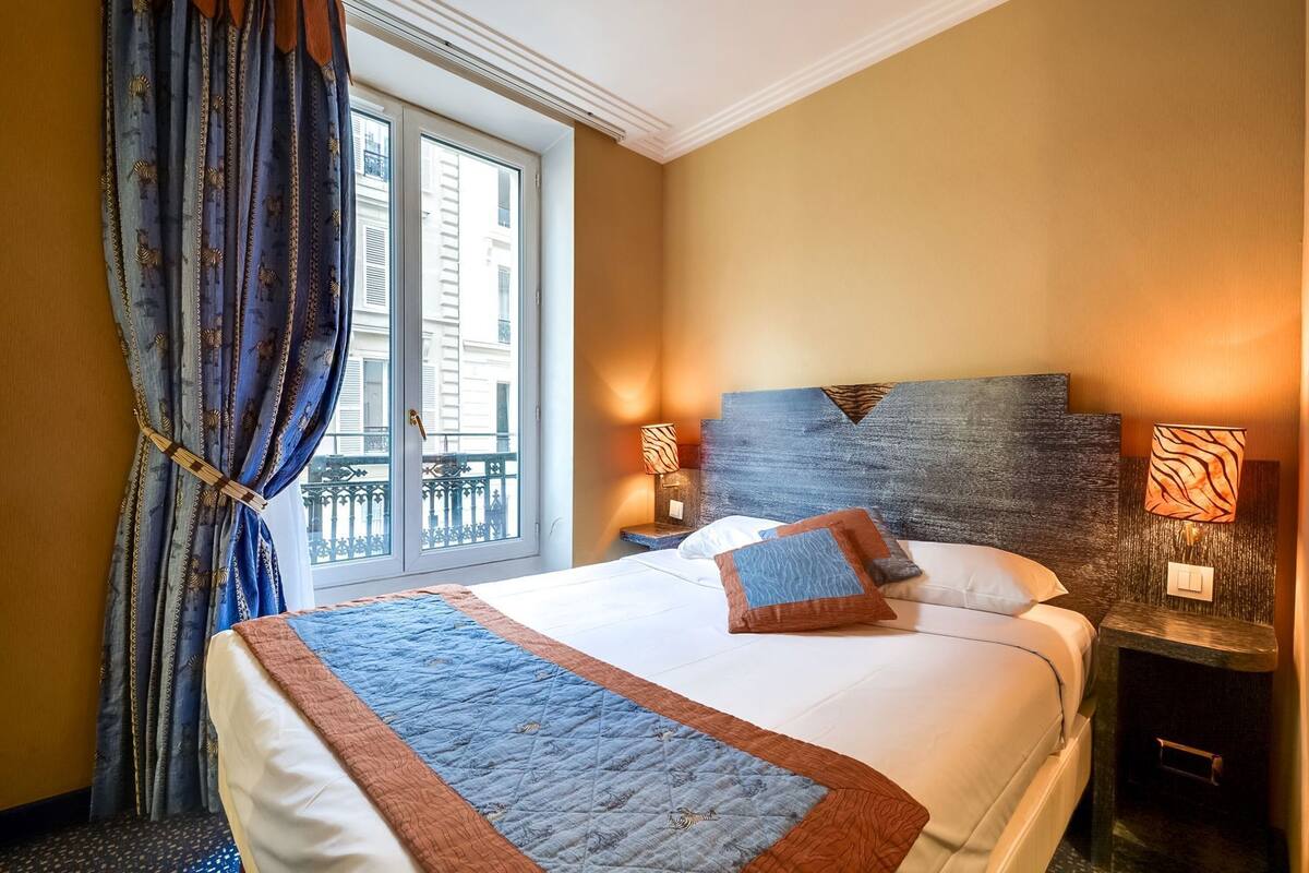 A hotel room with a large bed, a view through a window, and a nightstand with a lamp.