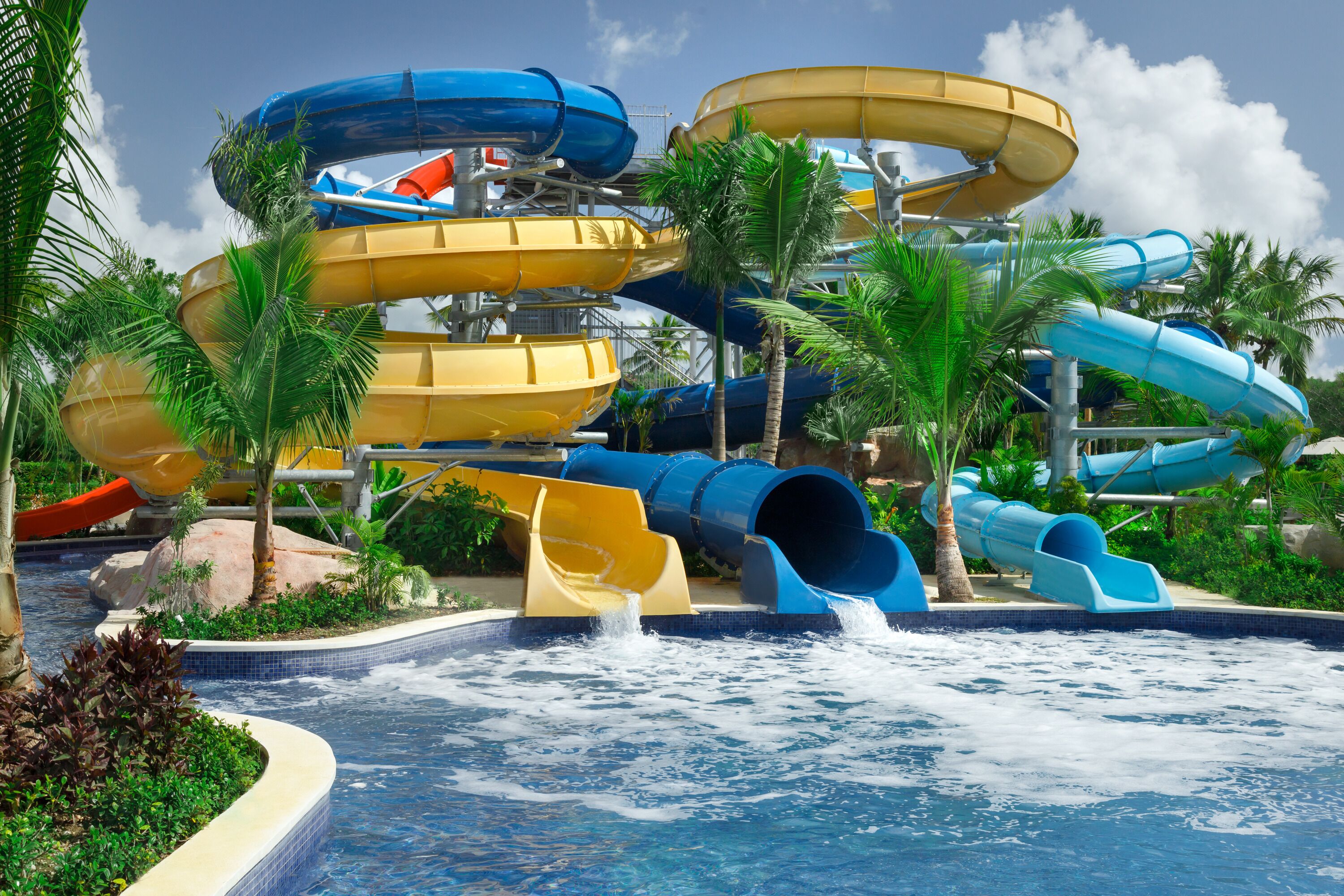 Een waterpark met diverse glijbanen in blauw, geel en rood, omringd door palmbomen en een zwembad.