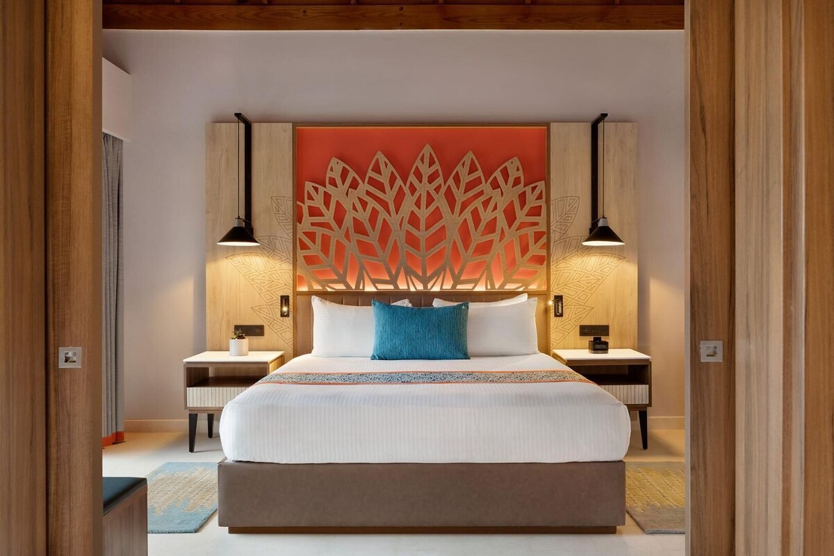 A modern hotel room with a large bed, wooden headboard, two bedside tables with lamps, and a decorative wall panel.