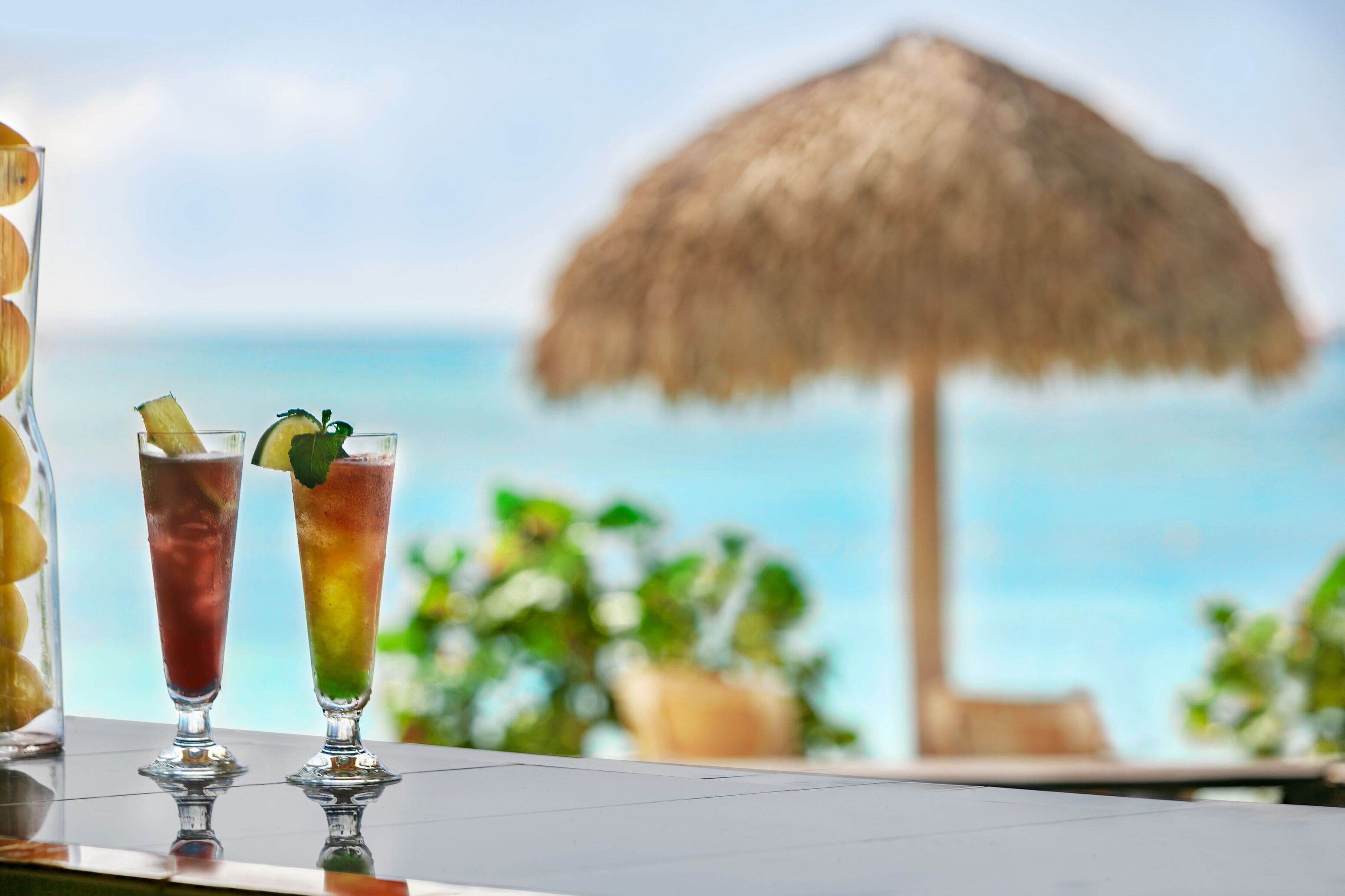 Zwei Cocktails auf einem Tisch mit Blick auf das Meer und einem Strohdachschirm im Hintergrund.