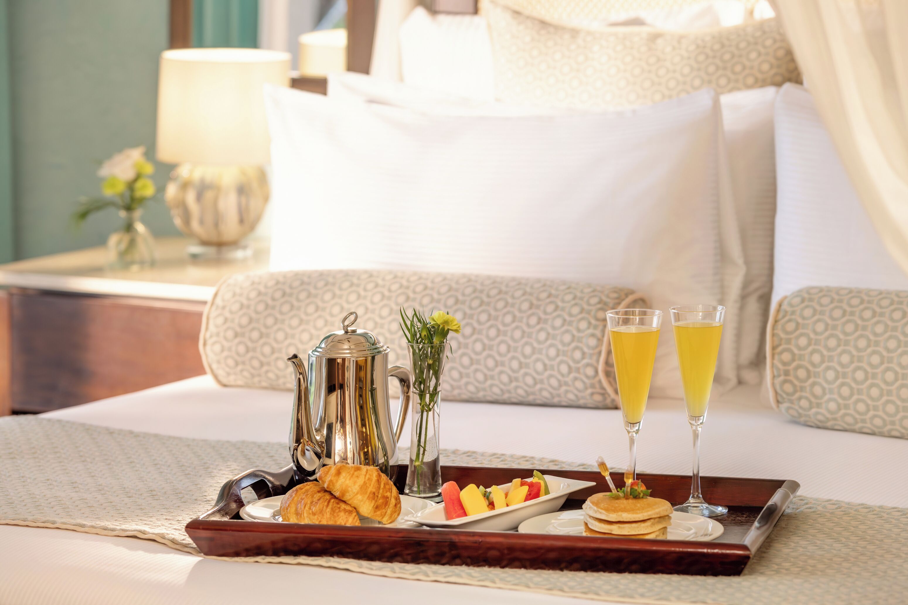 A breakfast tray with croissants, fruit, pancakes, and two glasses of orange juice on a bed with a patterned pillow and a bedside table with a lamp and a vase of flowers.