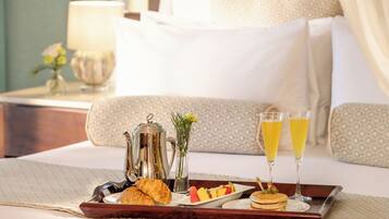 A breakfast tray with croissants, fruit, pancakes, and two glasses of orange juice on a bed with a patterned pillow and a bedside table with a lamp and a vase of flowers.