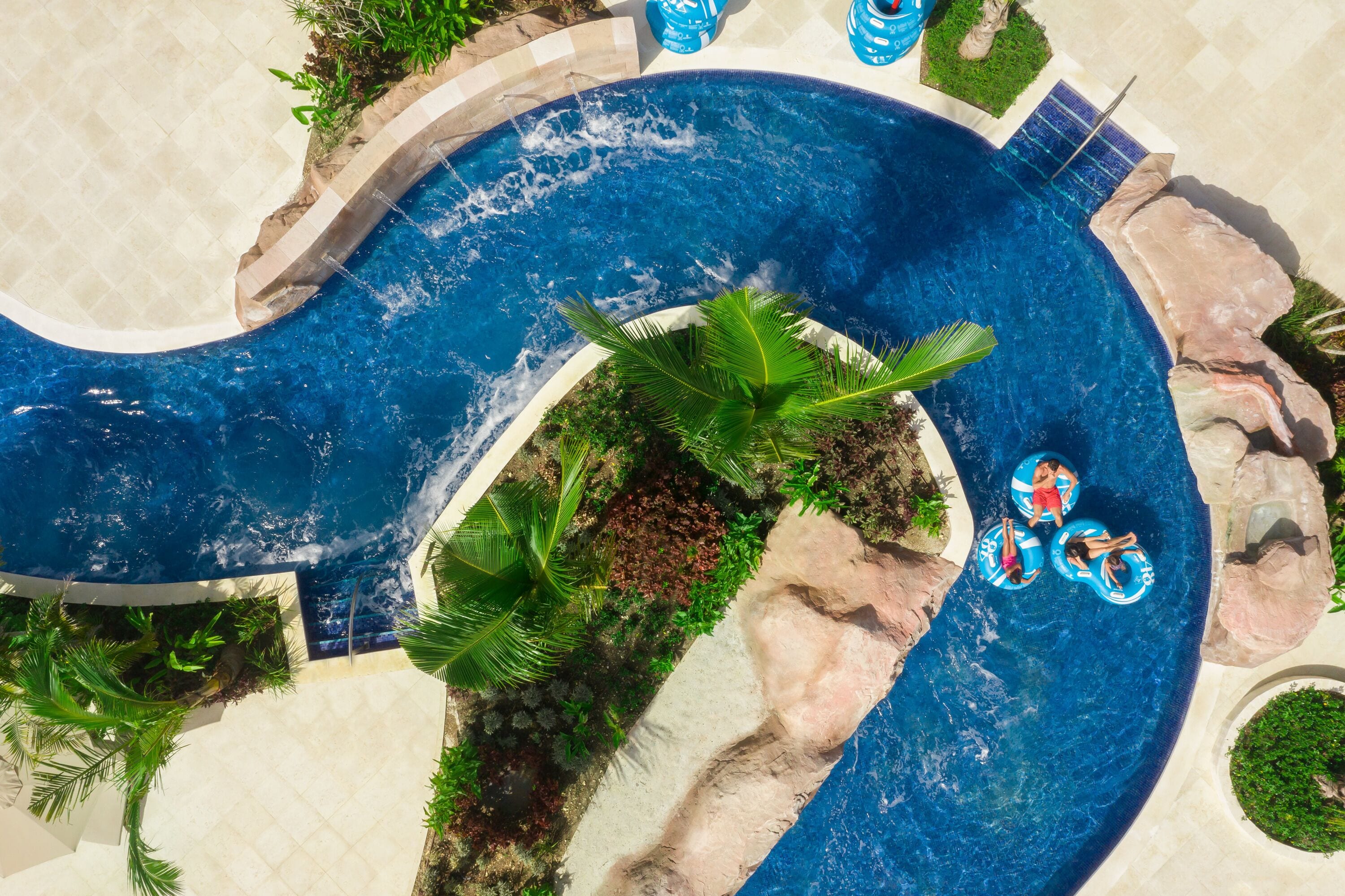 Una piscina grande y serpenteante con una isla central, rodeada de plantas tropicales y rocas.