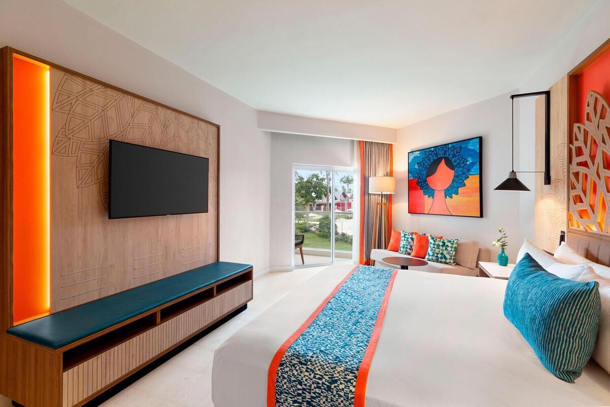 A modern hotel room with a large bed, a TV mounted on a wooden panel, a sofa, a small table, and a view of the outdoors.