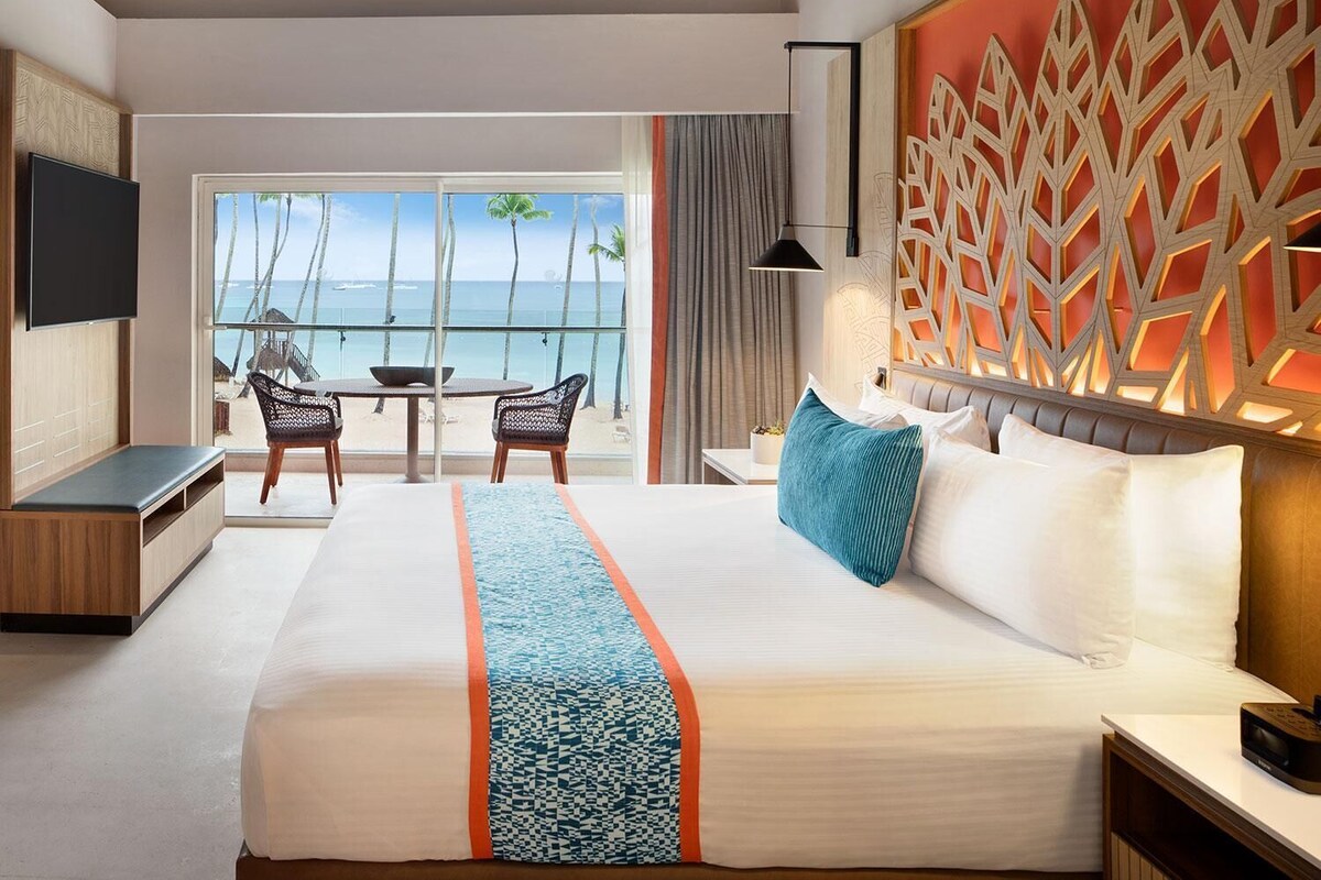A hotel room with a large bed, a TV, a balcony with a view of the sea, and a dining area.