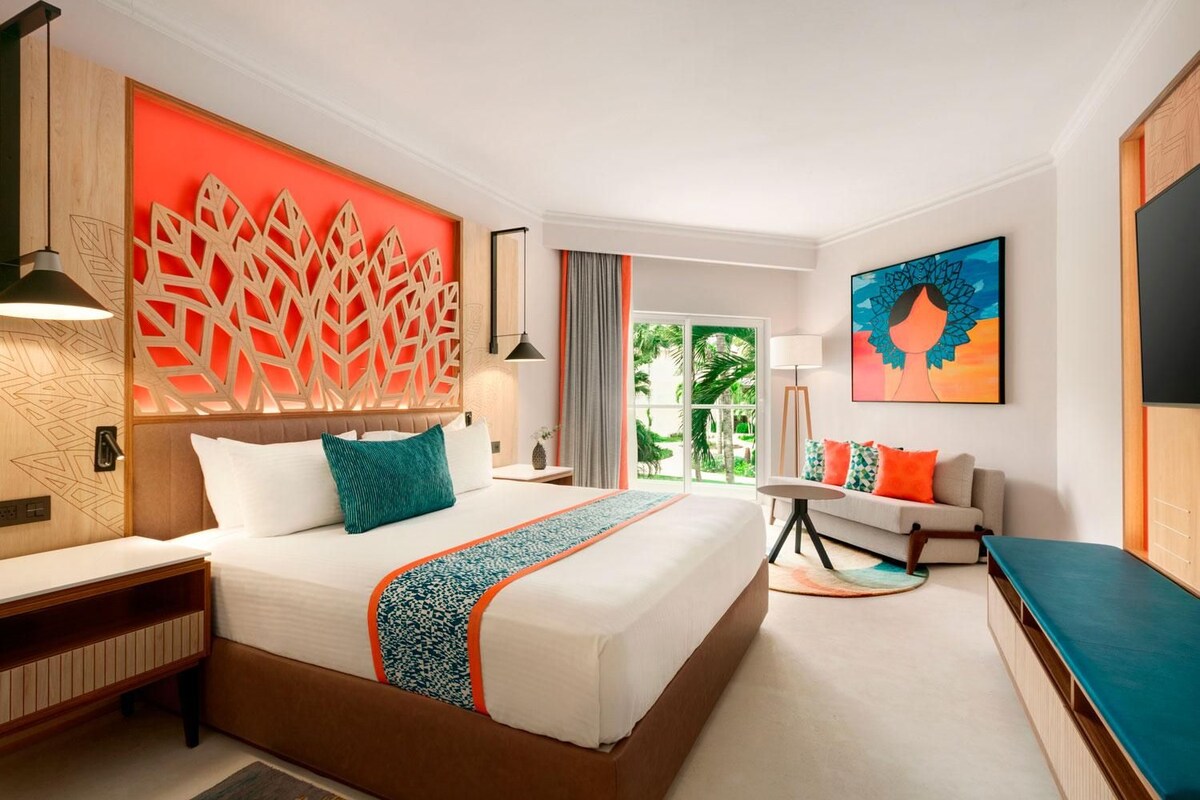 A hotel room with a large bed, a sofa, a TV, and a balcony with a view of palm trees.