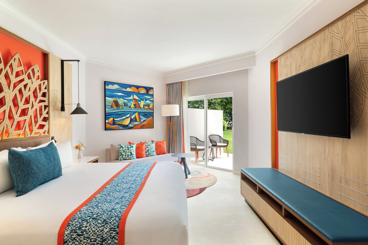 A modern hotel room with a large bed, a flat-screen TV, a seating area, and a view of the outdoors.