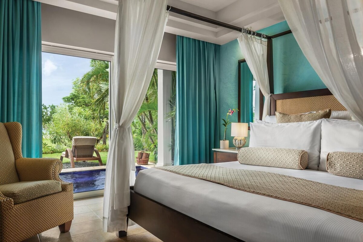 A spacious bedroom with a large bed, a sitting area with a chair, a nightstand with a lamp, and a view of a swimming pool and greenery outside.