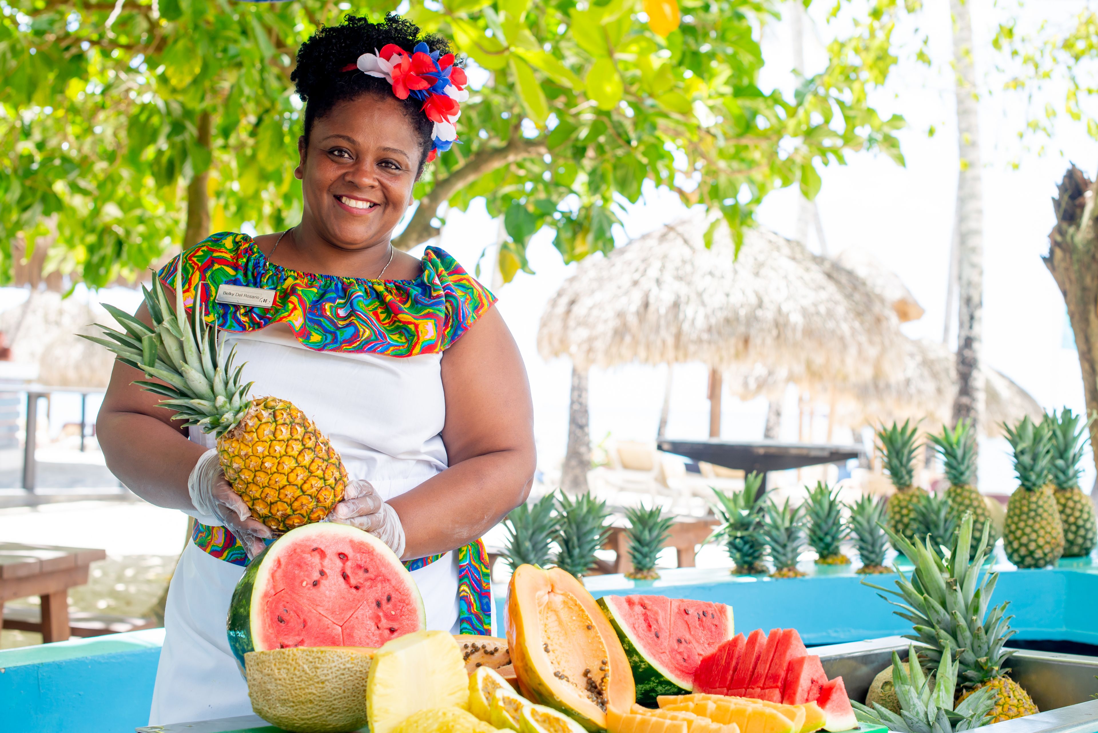 Una persona sosteniendo una piña y una sandía, con una mesa llena de diversas frutas en un entorno tropical.