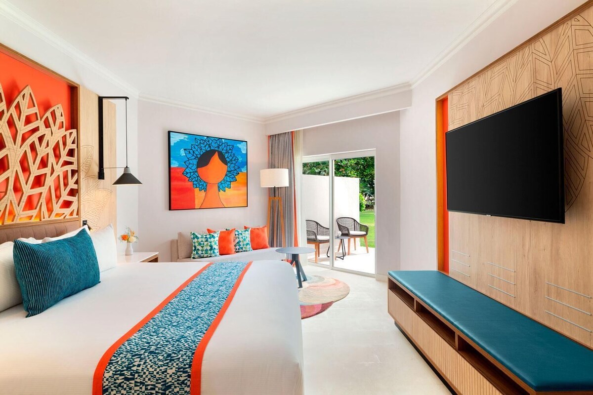 A hotel room with a large bed, a TV, a painting, and a view of the outdoors.