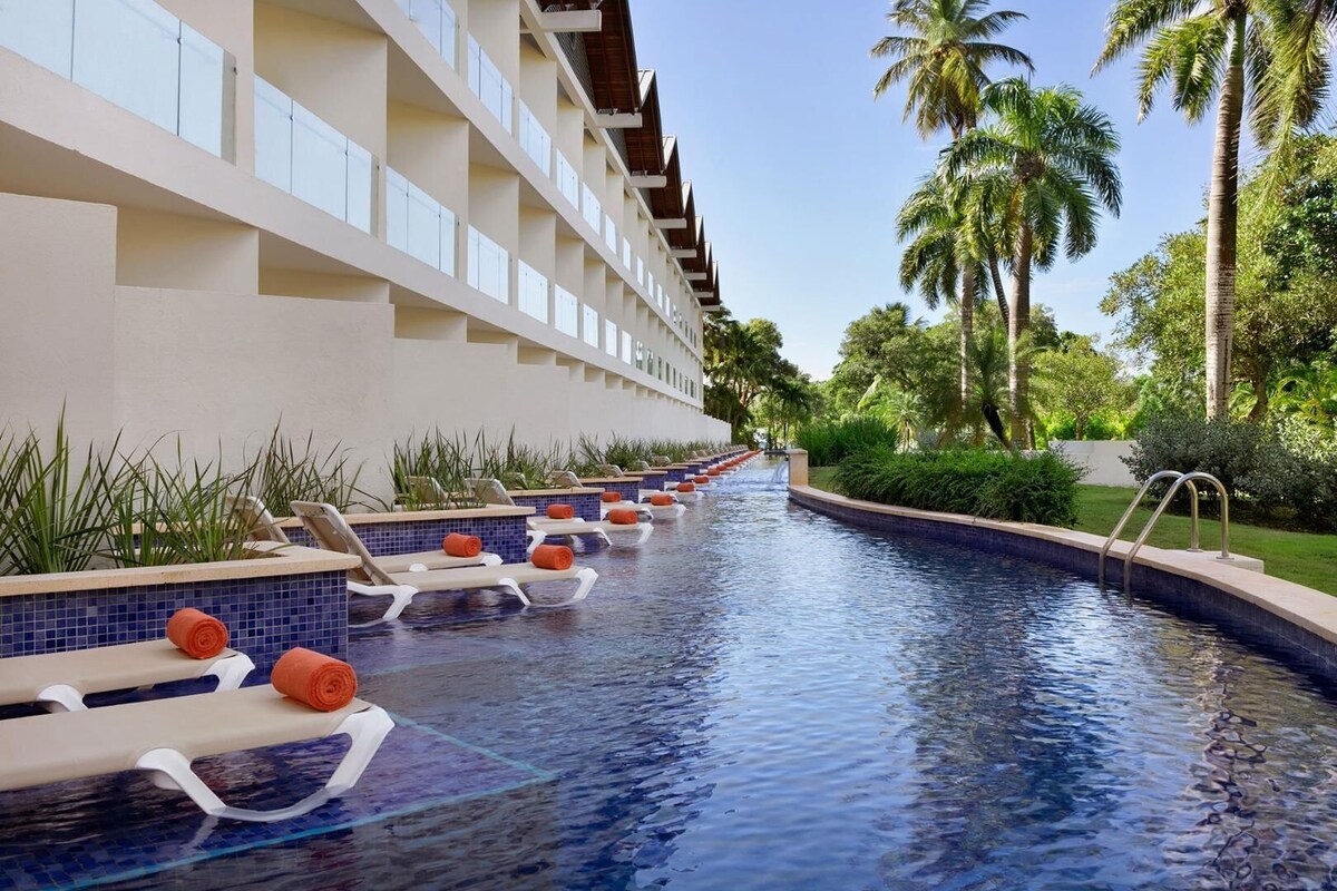 A pool area with lounge chairs and a clear blue pool, surrounded by palm trees and a building with balconies.