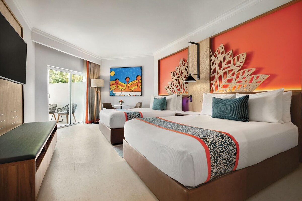 A hotel room with a large bed, a smaller bed, a desk, and a chair. There is a painting on the wall and a decorative wall panel.