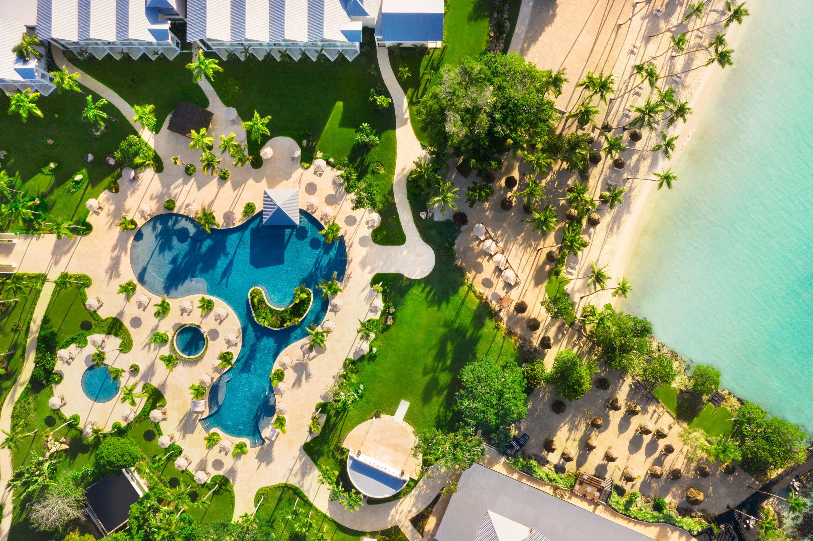 Vista aérea de un complejo turístico con múltiples piscinas, una zona de playa con tumbonas y una vista despejada del océano.