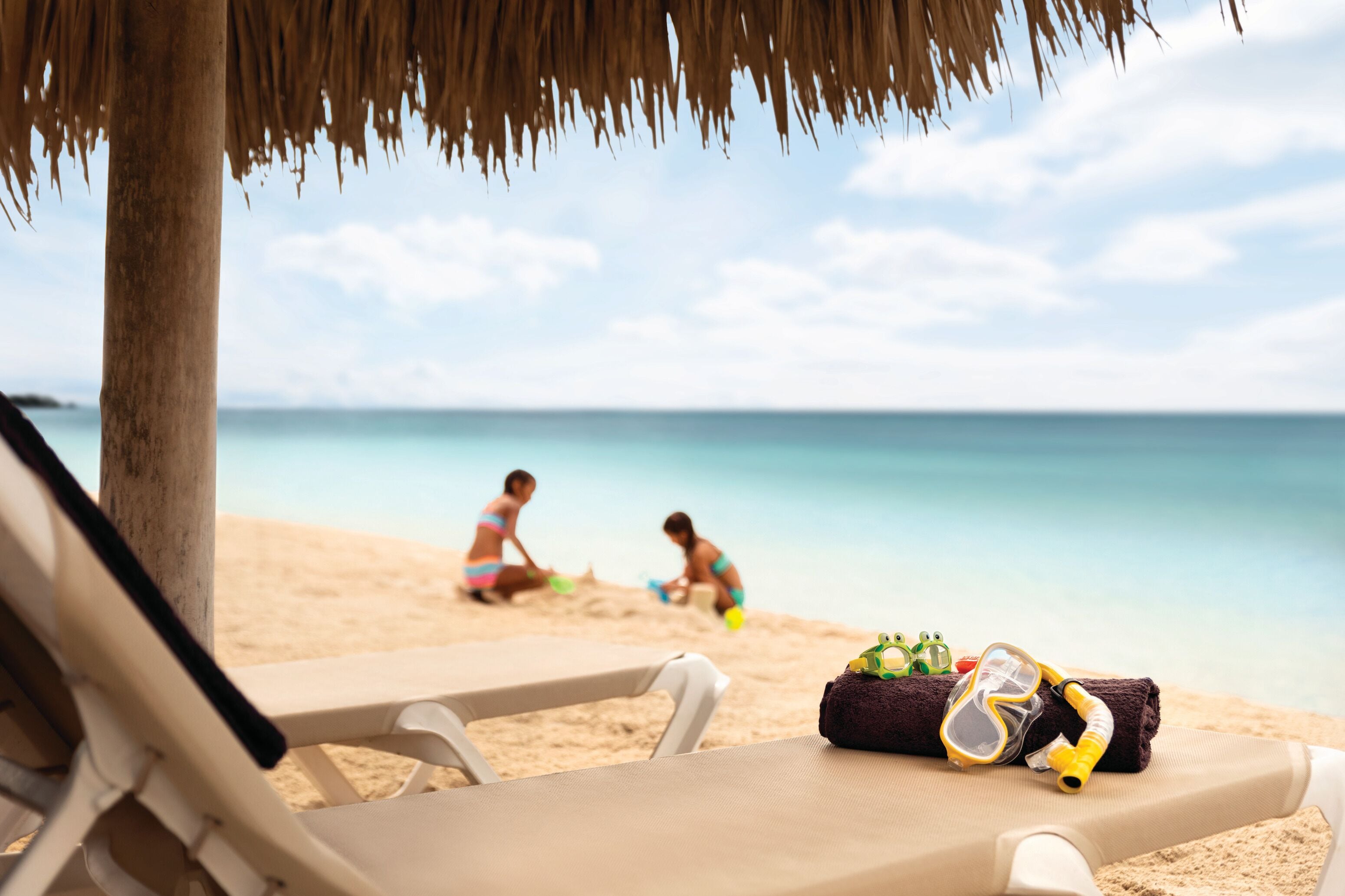 A beach scene with a thatched umbrella, a lounge chair, and beach toys on the sand.