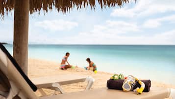 A beach scene with a thatched umbrella, a lounge chair, and beach toys on the sand.