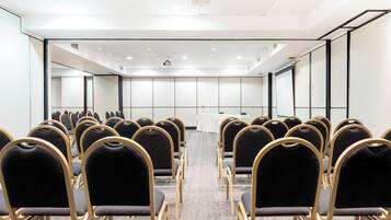 Un salón de conferencias con filas de sillas, un proyector y una mesa blanca con una planta.