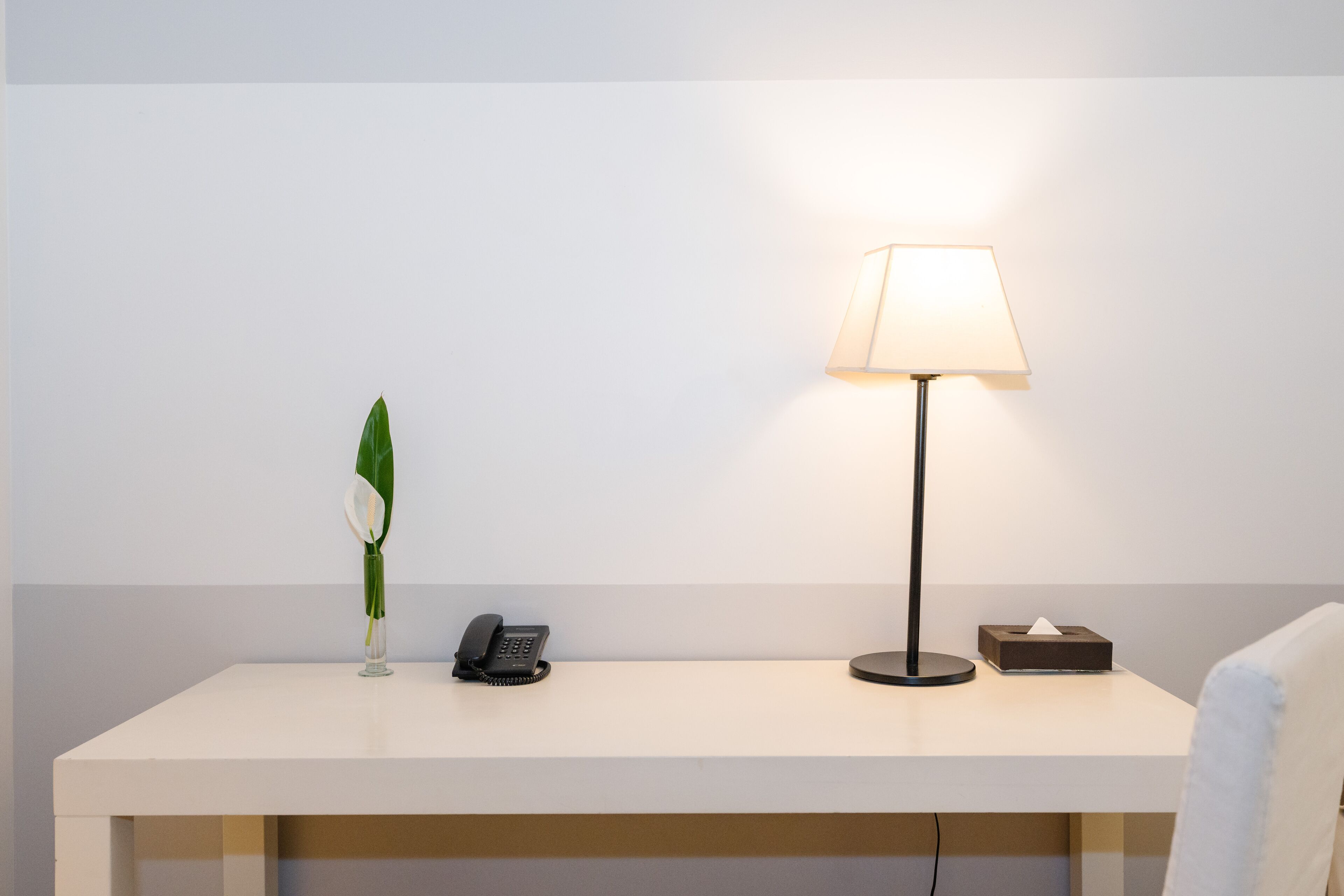A clean, minimalist desk with a lamp, a telephone, a tissue box, and a vase with a single flower.