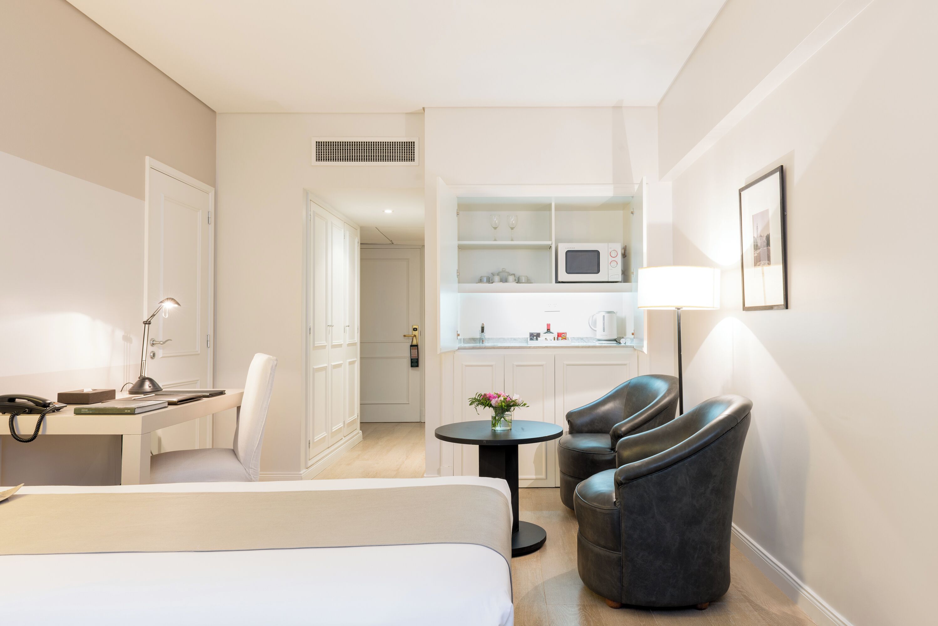 A hotel room with a bed, desk, chair, and a small table with a lamp. There is a kitchenette with a microwave and a coffee maker.