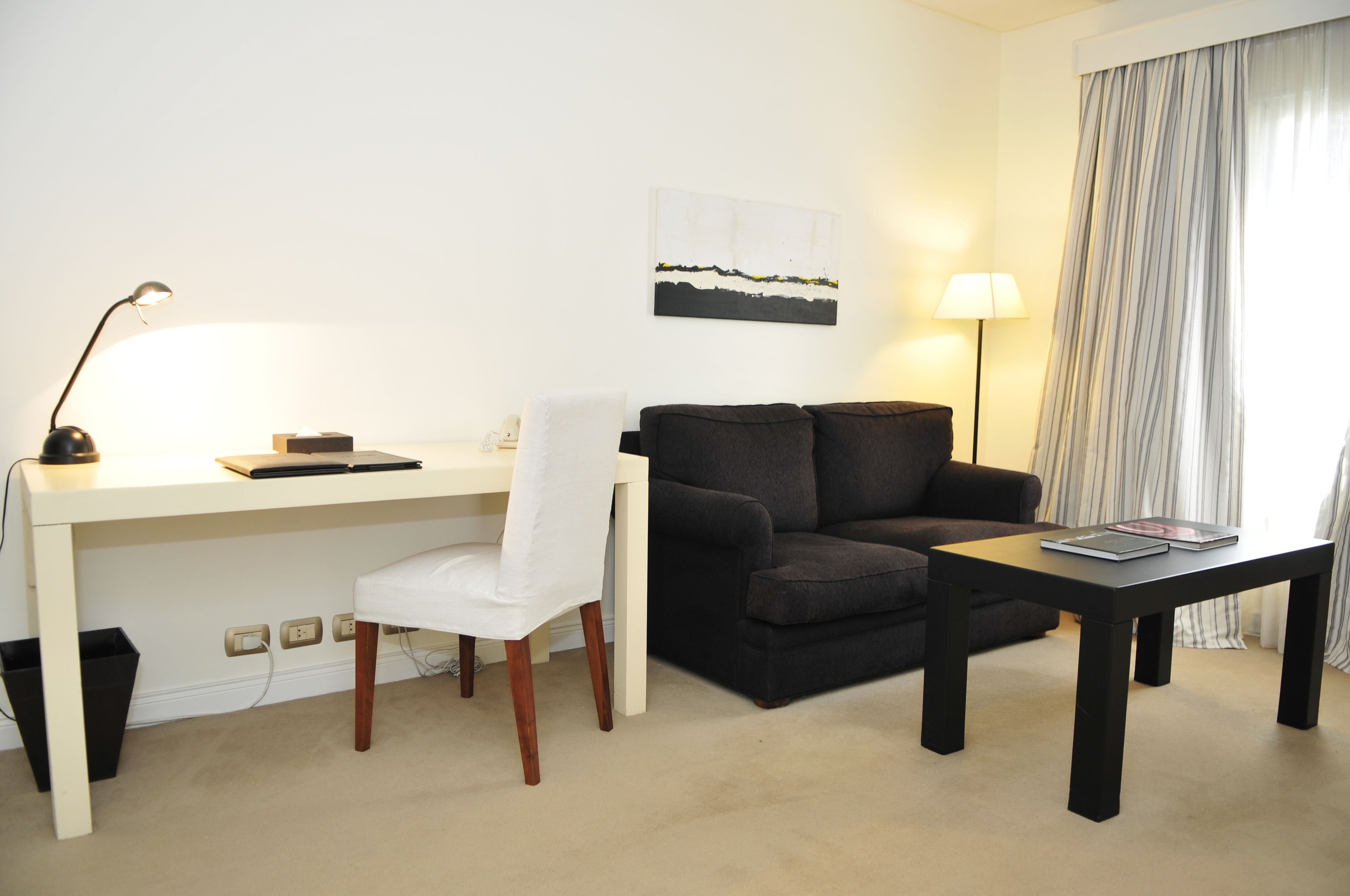 Habitació d'hotel amb un escriptori blanc, un sofà negre, una taula petita i una làmpada.
