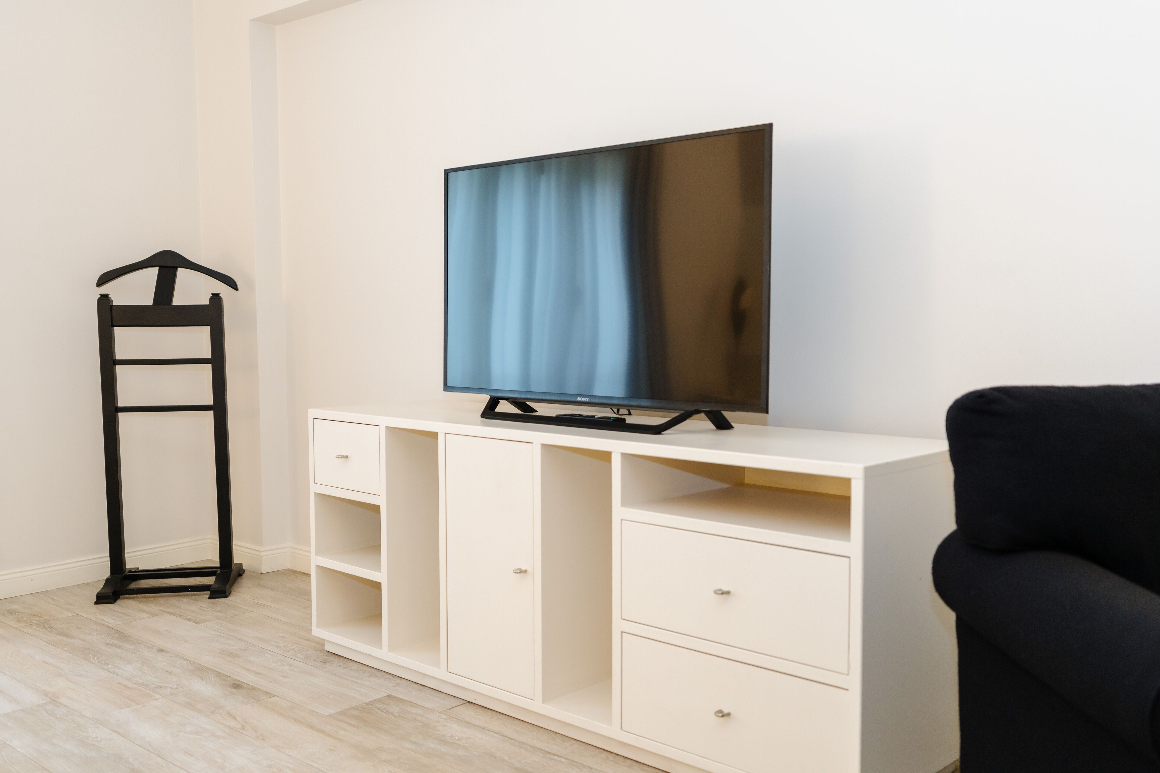 Una habitación con televisión sobre un mueble blanco, una silla negra y un perchero.