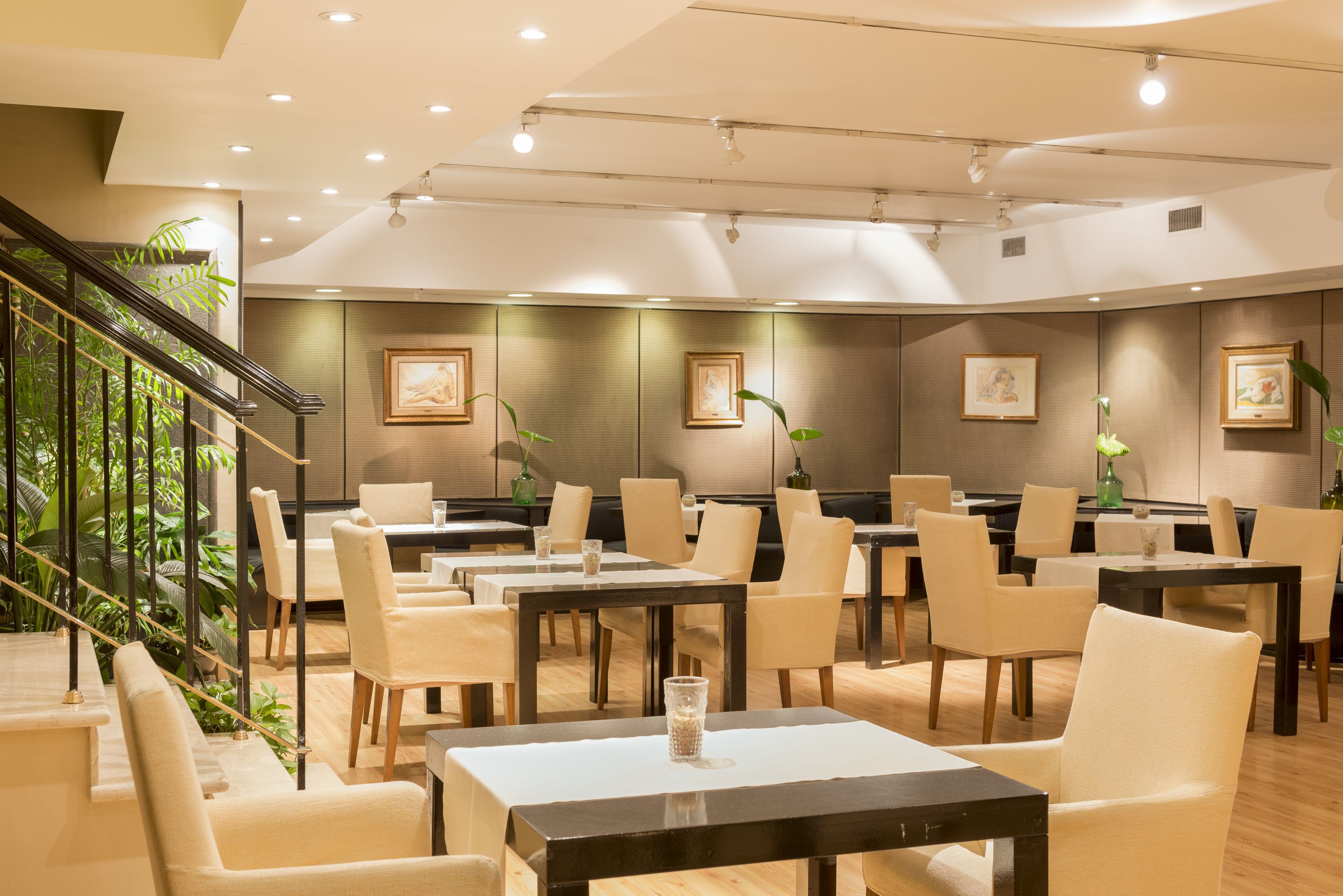 A spacious restaurant with high ceilings, recessed lighting, and a staircase with plants. Tables are arranged for dining, and artwork is displayed on the walls.