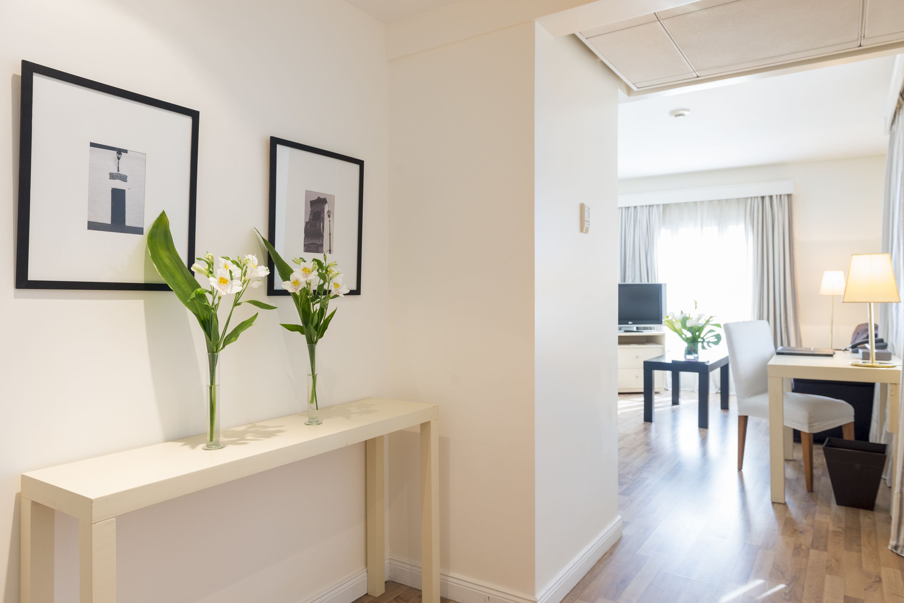 Habitación de hotel con una mesa consola blanca, dos cuadros enmarcados, un escritorio con silla, un televisor y una lámpara.