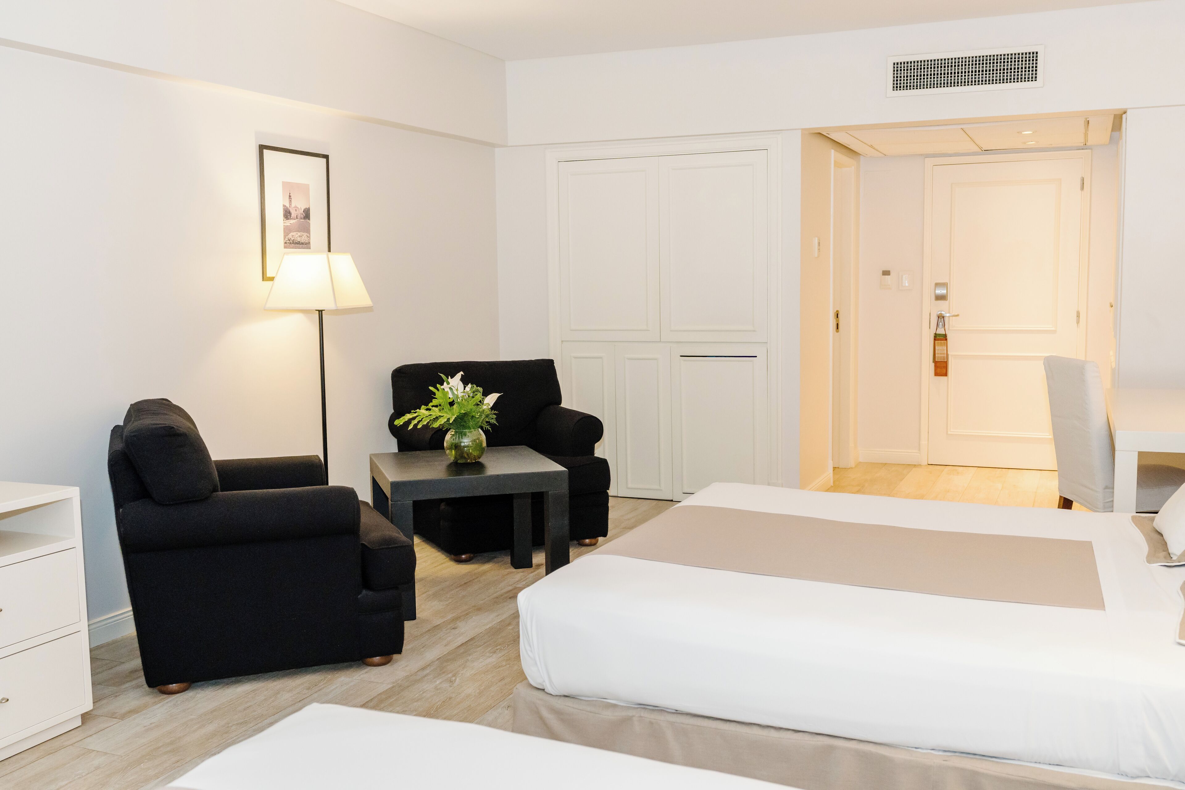 A hotel room with a bed, a sofa, a small table with a lamp, and a door leading to another room.