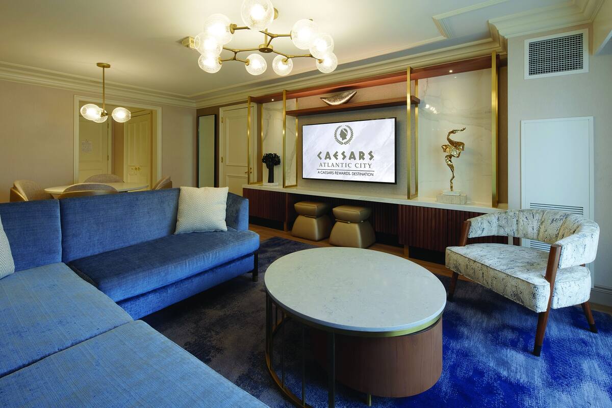 Ein modernes Wohnzimmer mit einem blauen Modulsessel, einem runden Couchtisch und einem klassischen Sessel. Ein großer Bildschirm zeigt das Logo von Caesars Atlantic City.