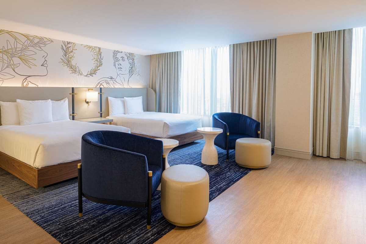 Ein Hotelzimmer mit zwei Betten, einem blauen Sessel, einem runden Hocker, einem kleinen Tisch und einer Wanddekoration.