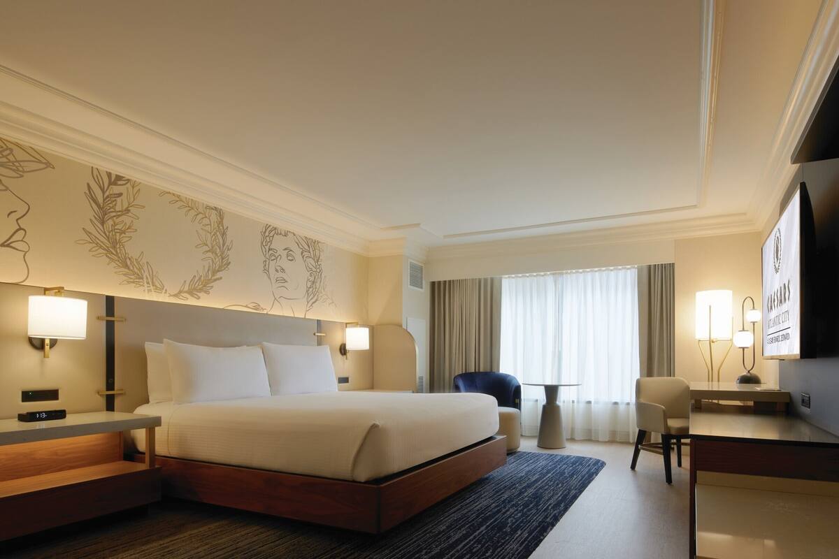 Ein Hotelzimmer mit einem großen Bett, Nachttischen, einem Schreibtisch, einem Sessel und einem Fernseher, sowie einer Wand mit Kunstwerken.