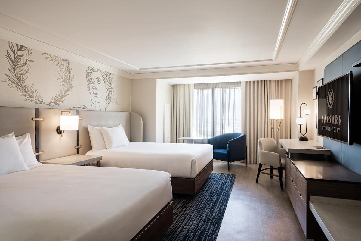 Ein Hotelzimmer mit zwei Betten, einem Schreibtisch, einem Sessel, einem Fernseher und einem Fenster mit Vorhängen.