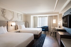 Ein Hotelzimmer mit zwei Betten, einem Schreibtisch, einem Sessel, einem Fernseher und einem Fenster mit Vorhängen.