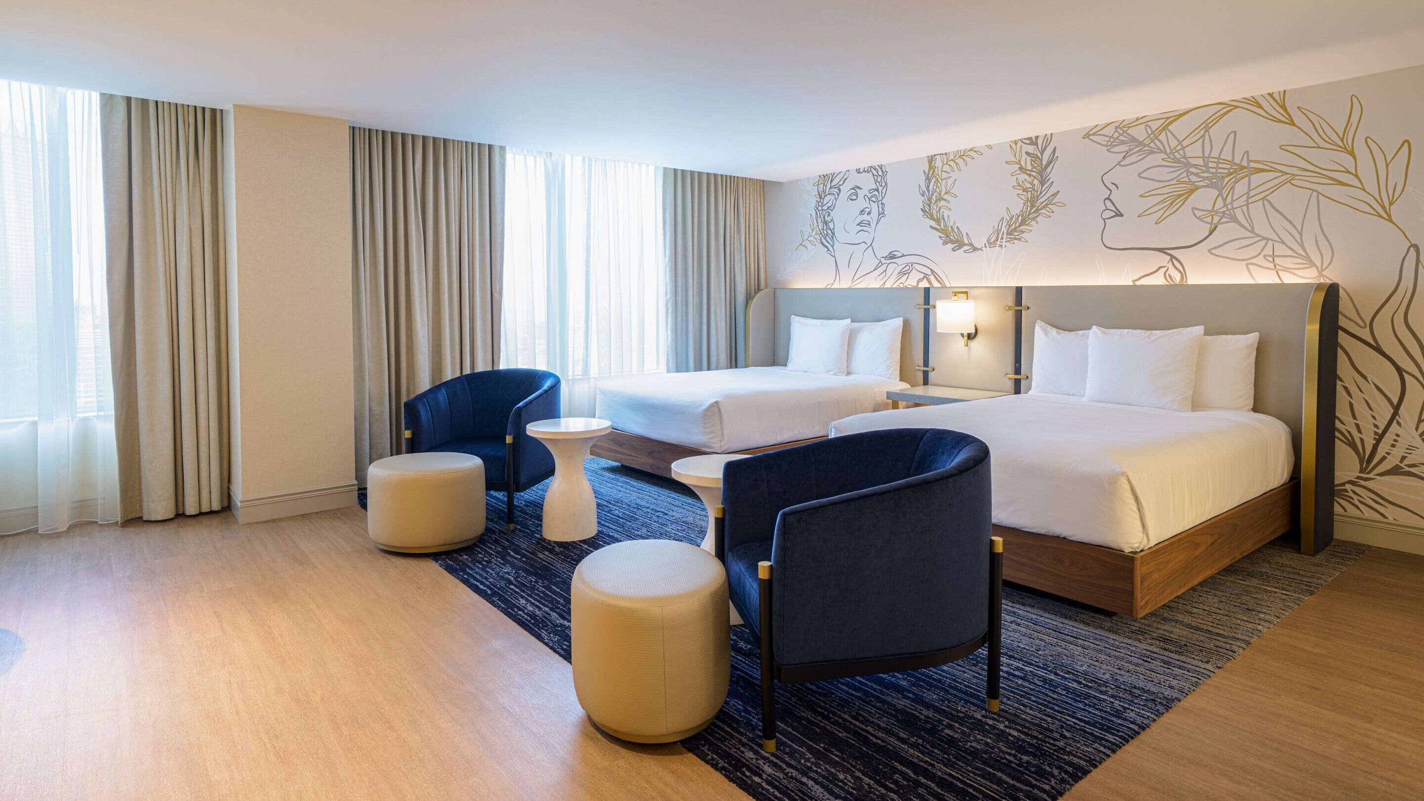 A hotel room with two beds, a blue armchair, a round ottoman, and a small table.
